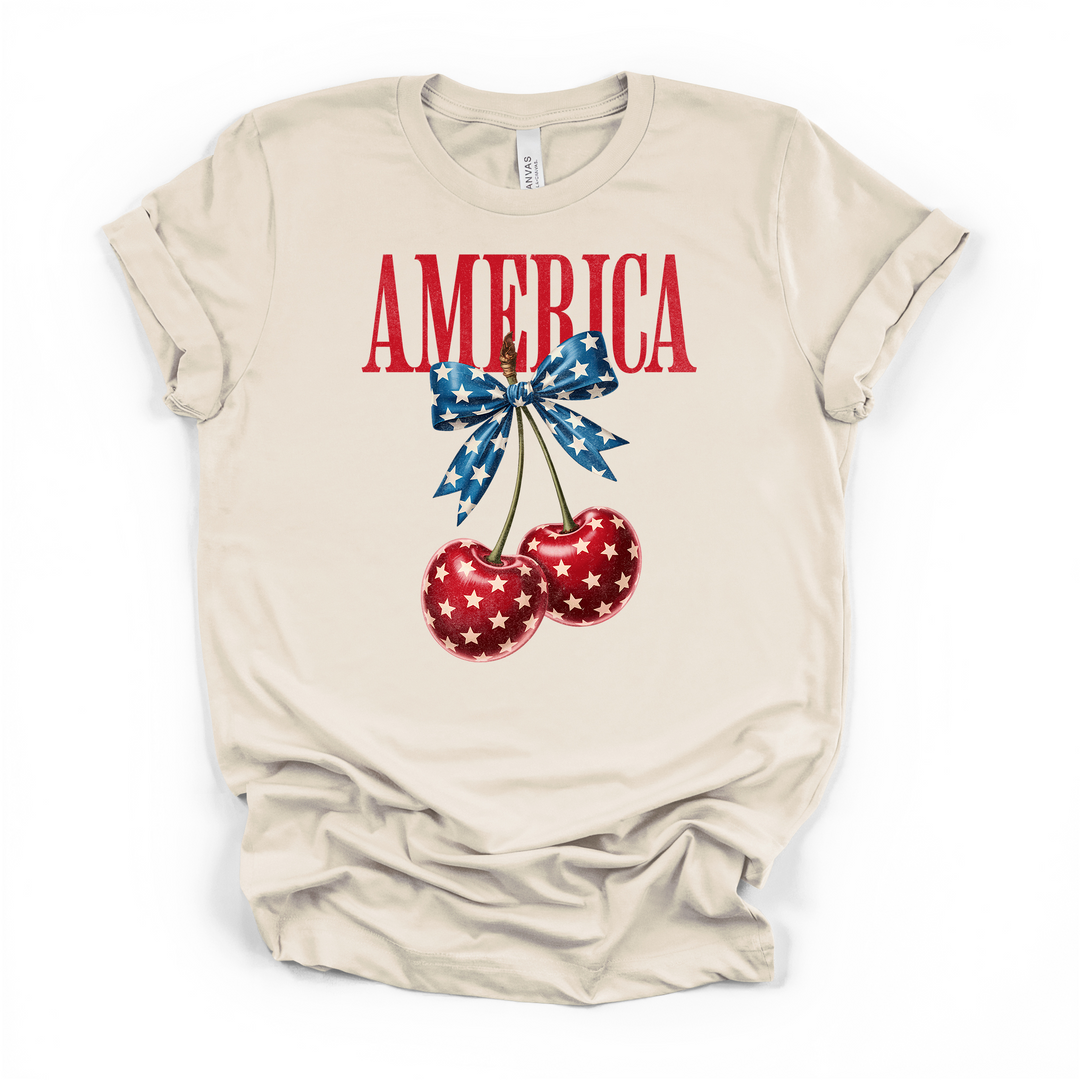 Natural cream t-shirt with two red star-covered cherries tied with a blue star bow graphic and bold red America text printed on the front