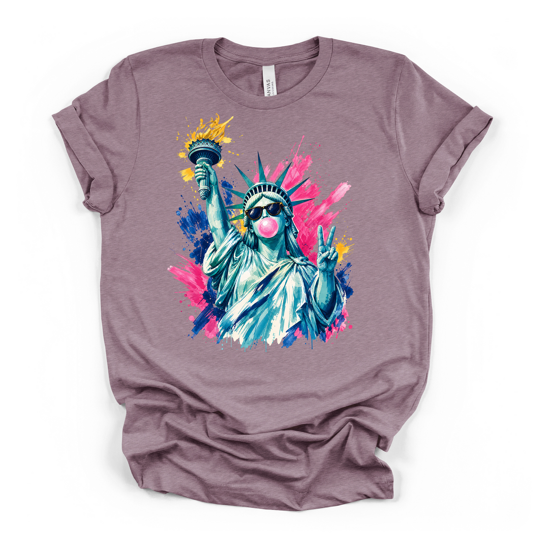 Heather purple t-shirt with a colorful watercolor Statue of Liberty in sunglasses blowing a pink bubble and throwing a peace sign on a bold paint splash graphic
