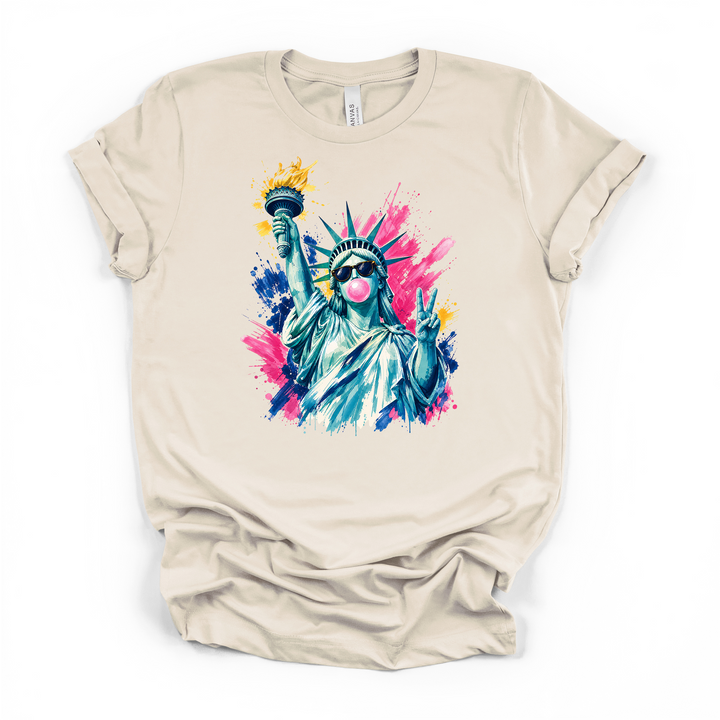 Natural cream t-shirt with a bold pop-art Statue of Liberty graphic wearing sunglasses blowing bubble gum and giving a peace sign surrounded by pink blue and yellow paint splashes