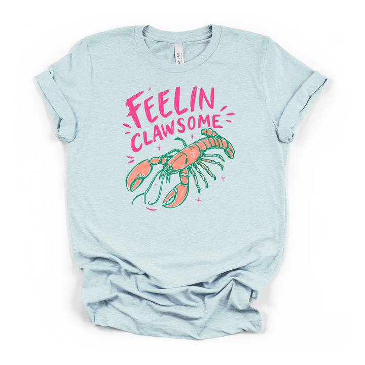 Heather ice blue t-shirt with a bold illustrated lobster in coral and green with pink sparkles and Feelin Clawsome printed in hot pink text on the front