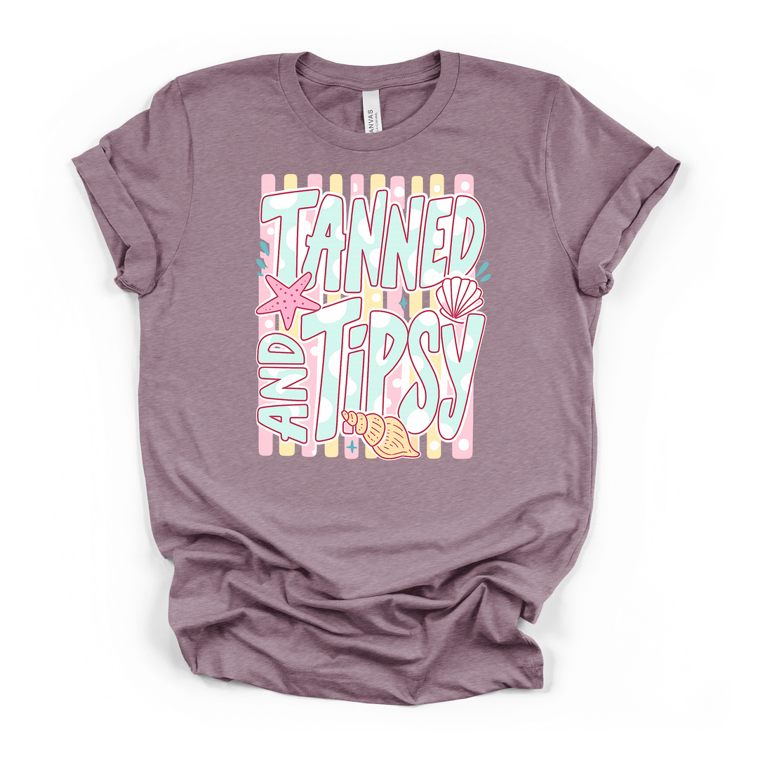 Heather purple t-shirt with a pastel Tanned and Tipsy graphic featuring bold retro lettering a starfish scallop shell and conch on a striped background