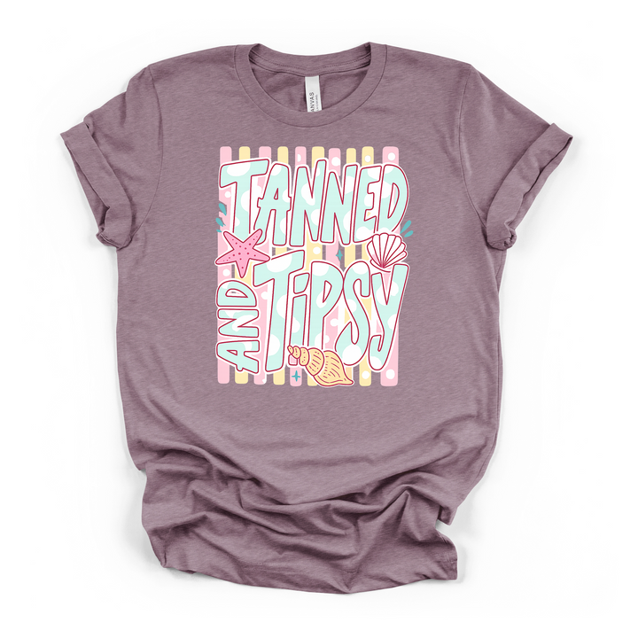 Heather purple t-shirt with a pastel Tanned and Tipsy graphic featuring bold retro lettering a starfish scallop shell and conch on a striped background
