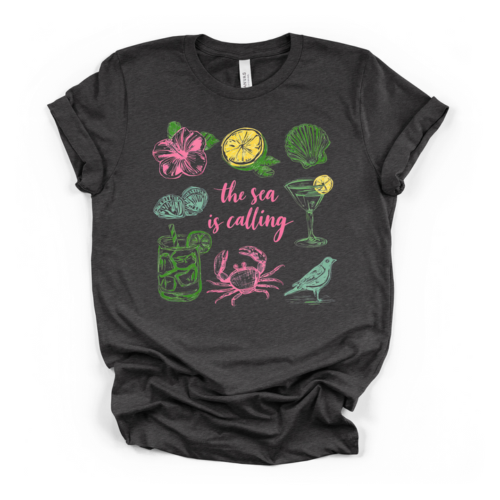 The Sea is Calling beach graphic t-shirt in dark grey with colorful coastal icons including crab, seashell, hibiscus, and cocktails
