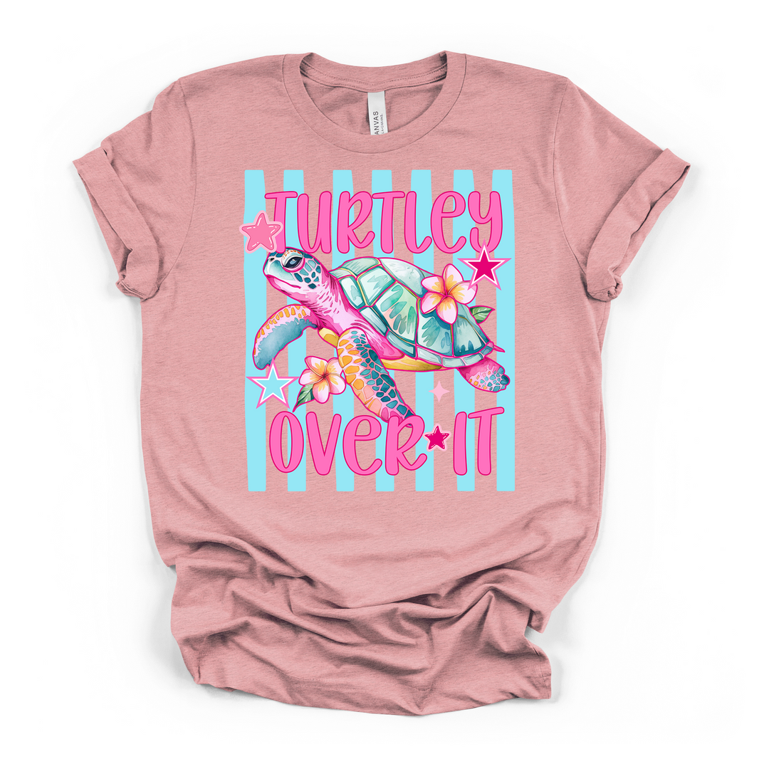 Heather pink t-shirt featuring a bright colorful sea turtle with tropical flowers and stars on a blue striped background and Turtley Over It in pink text