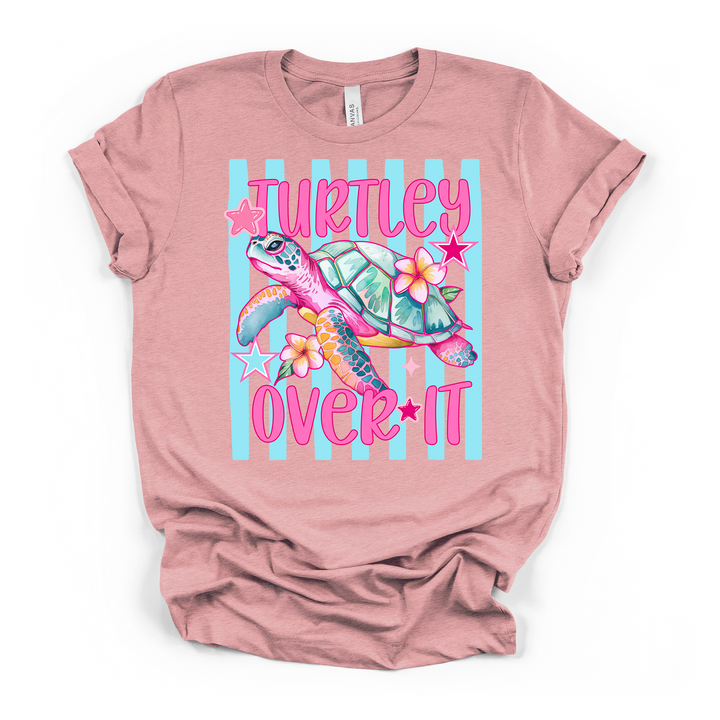 Heather pink t-shirt featuring a bright colorful sea turtle with tropical flowers and stars on a blue striped background and Turtley Over It in pink text