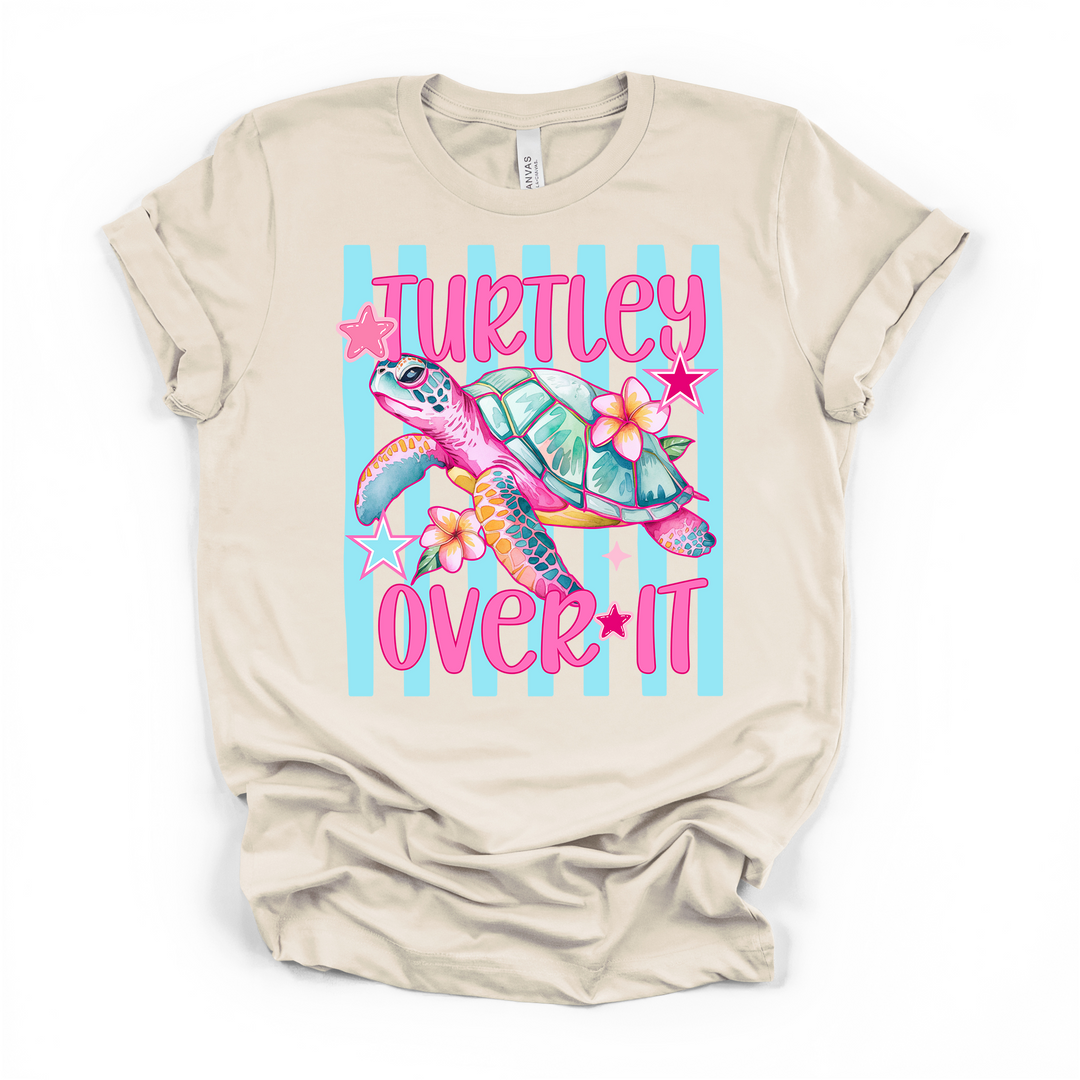 Natural cream t-shirt with a colorful pink sea turtle and plumeria flowers on a blue striped background with Turtley Over It printed in hot pink